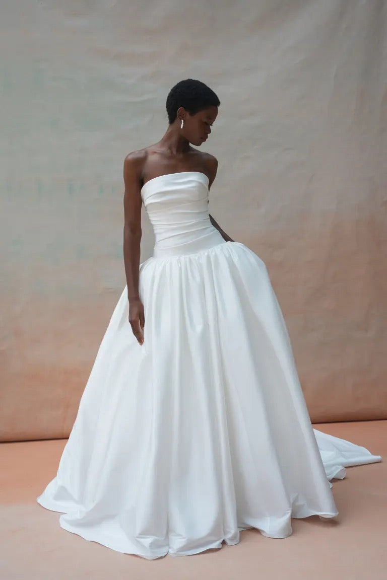 A person adorned in the opulent Charlotte Wedding Dress by Jenny Yoo from Bergamot Bridal, made from luxurious taffeta fabric, stands on a light-colored surface, gently touching the gathered ball skirt. The backdrop features a muted gradient texture.