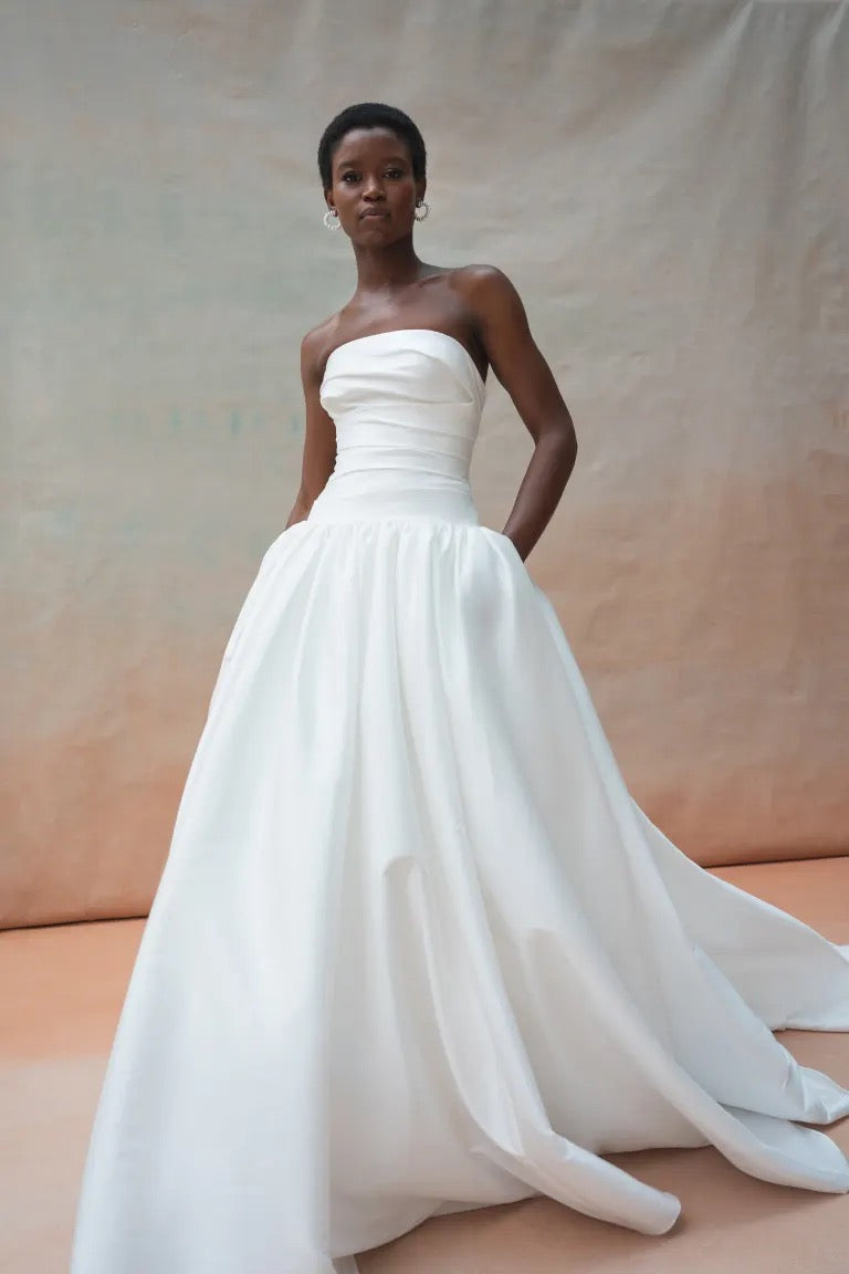 A person stands in the Charlotte Wedding Dress by Jenny Yoo, crafted from luxe taffeta fabric, complete with pockets and a gathered ball skirt from Bergamot Bridal, set against a neutral fabric background.