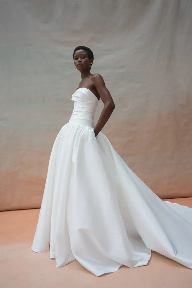 An individual stands in the Charlotte Wedding Dress by Jenny Yoo from Bergamot Bridal, crafted with luxe taffeta fabric. The gown features a strapless design and a full gathered ball skirt, with hands tucked casually into pockets, posed against a neutral backdrop.