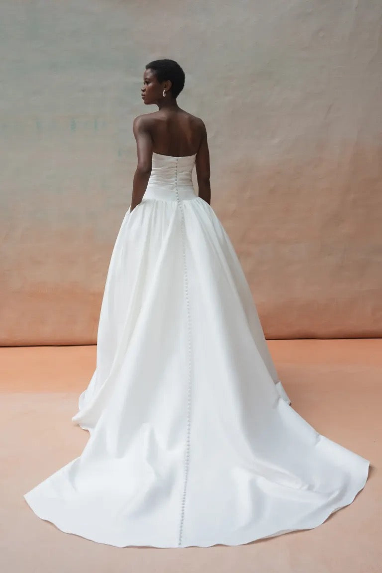 A person is shown from the back wearing a strapless white Charlotte Wedding Dress by Jenny Yoo, available at Bergamot Bridal. The gown boasts a gathered ball skirt made of luxe taffeta fabric and features a long train, all set against a neutral backdrop.