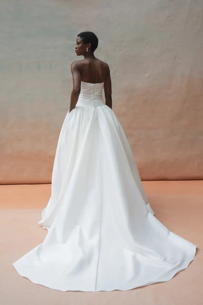 A person is shown from the back wearing a strapless white Charlotte Wedding Dress by Jenny Yoo, available at Bergamot Bridal. The gown boasts a gathered ball skirt made of luxe taffeta fabric and features a long train, all set against a neutral backdrop.