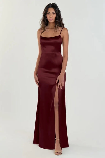 A woman posing in a sleek burgundy satin back crepe evening gown with a scoop neck and a thigh-high slit, wearing the Chase bridesmaid dress by Jenny Yoo from Bergamot Bridal.
