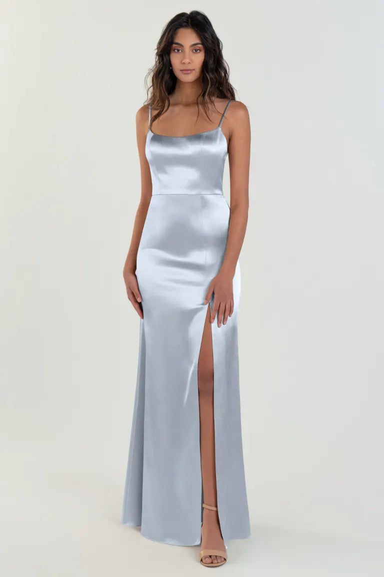 A woman stands in the Chase - Bridesmaid Dress by Jenny Yoo from Bergamot Bridal, made of silver satin back crepe with thin straps and a thigh-high slit, showcasing her legs. She complements the scoop neck bridesmaid dress, which features an elegant A-line skirt, with nude high-heeled sandals. The background is plain and light-colored.