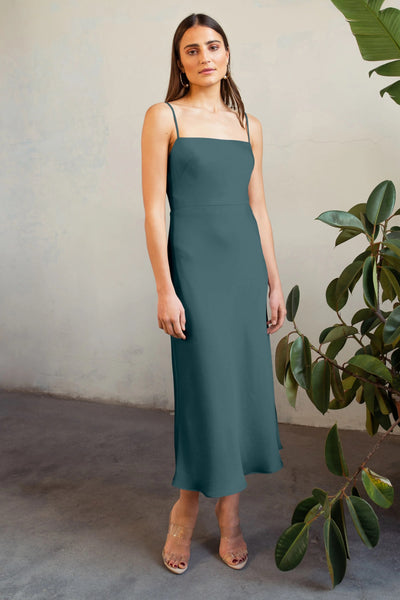 A woman in a Claudia satin midi dress by Jenny Yoo stands beside a potted plant, looking at the camera with a neutral expression.