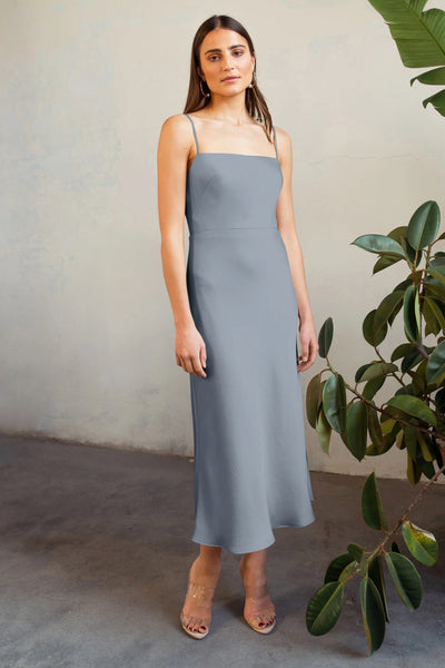 A woman wearing a gray Claudia - Bridesmaid Dress by Jenny Yoo and clear sandals stands in front of a textured wall with potted green plants nearby.