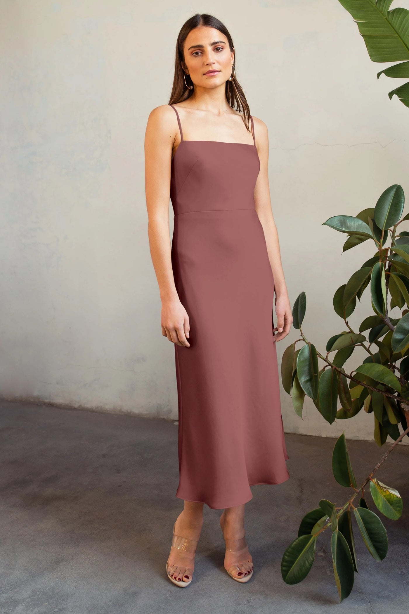 A woman in a Claudia - Bridesmaid Dress by Jenny Yoo in mauve satin midi dress stands confidently, posing beside a green potted plant from Bergamot Bridal.