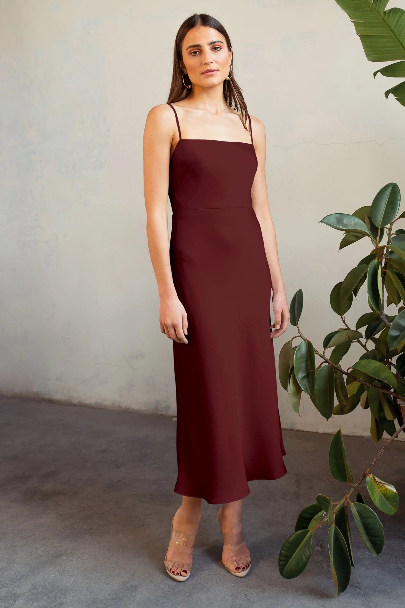 Woman in a burgundy satin midi Claudia - Bridesmaid Dress by Jenny Yoo standing by a plant with clear heeled sandals.