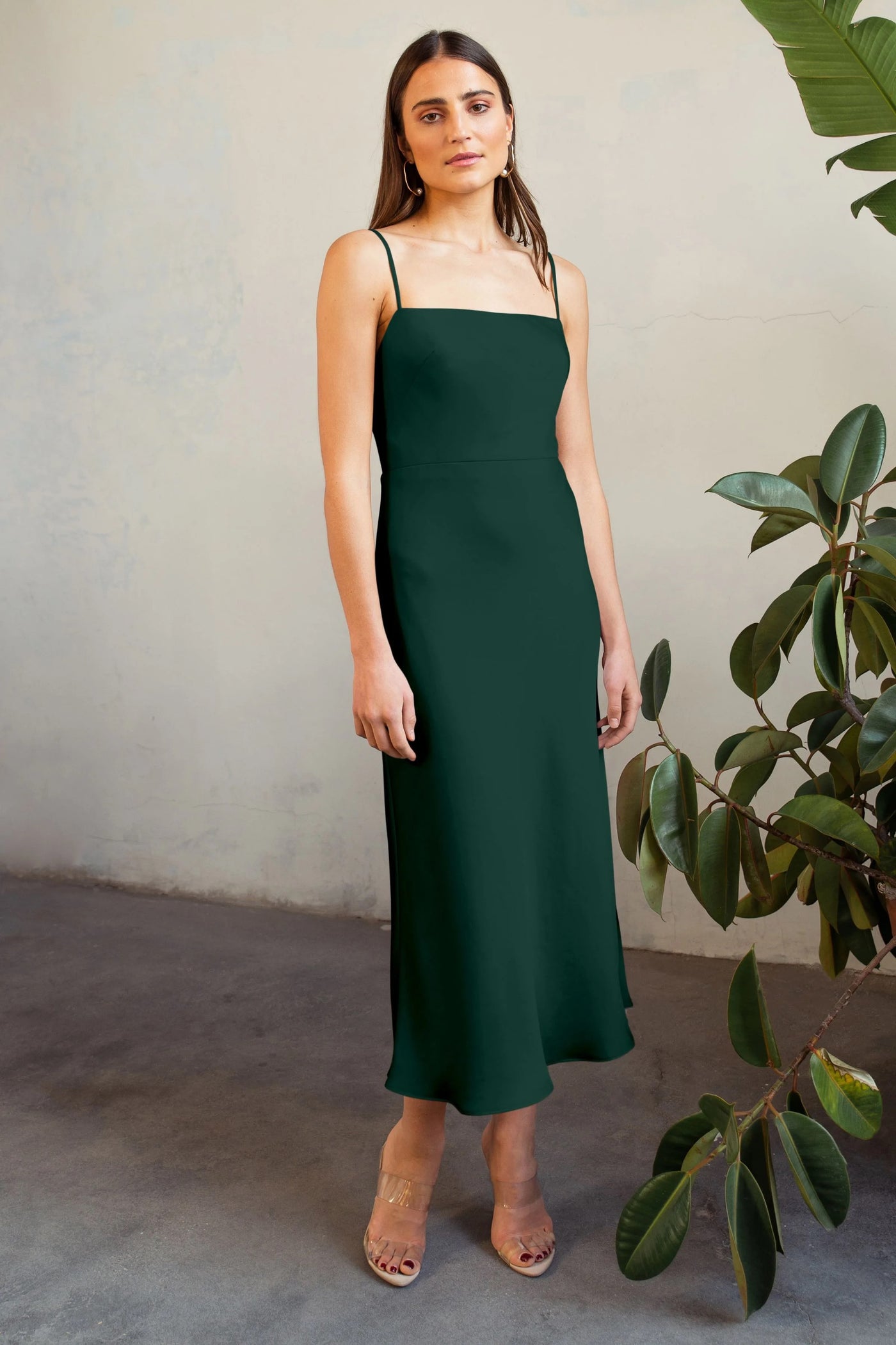 A woman in a dark green, sleeveless satin midi dress stands confidently near a plant, with neutral-toned footwear. (Claudia - Bridesmaid Dress by Jenny Yoo from Bergamot Bridal)