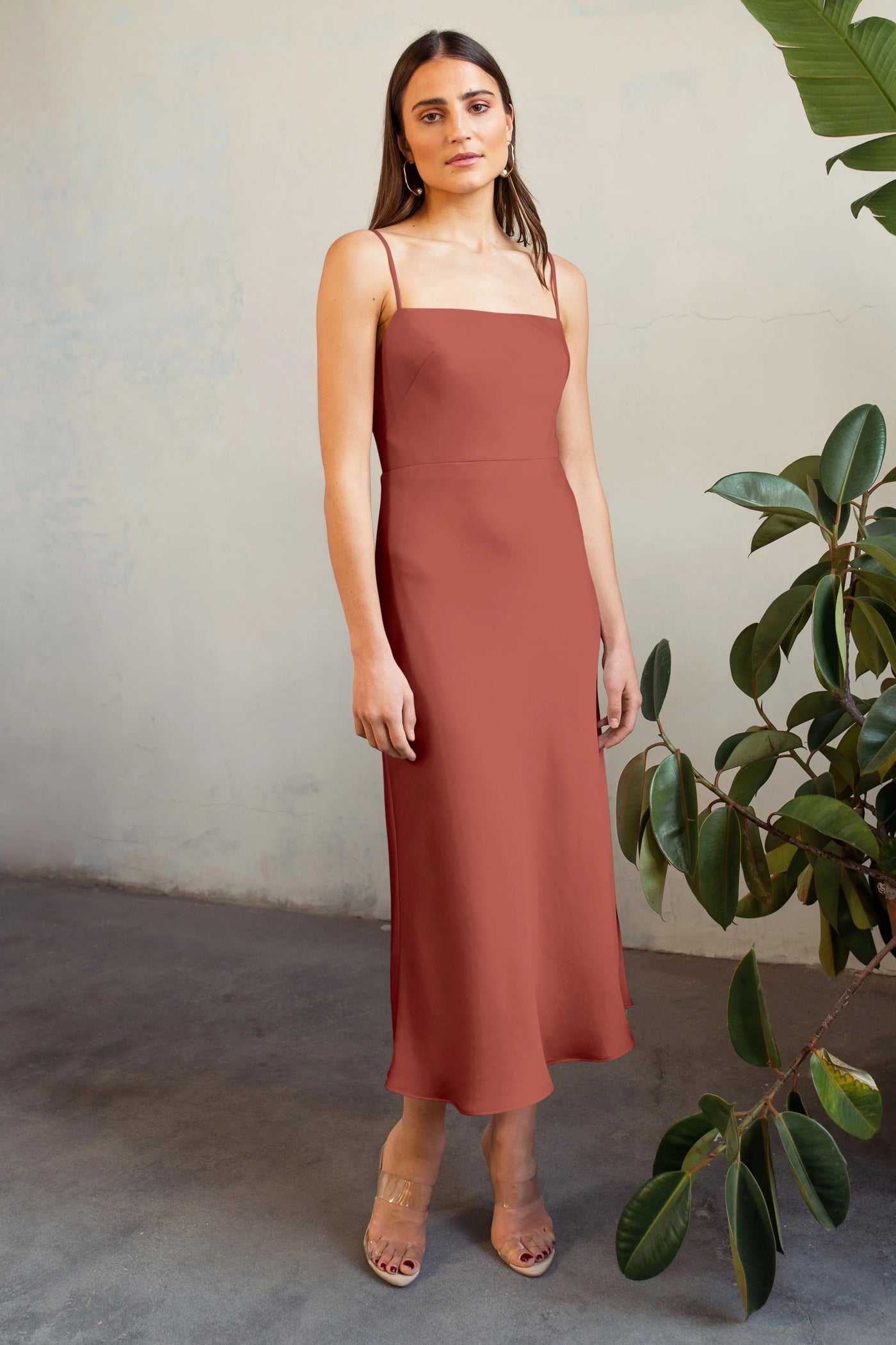 A woman in a chic terracotta satin midi dress stands against a textured wall near green foliage, wearing clear heeled sandals. The dress she is wearing is the Claudia Bridesmaid Dress by Jenny Yoo from Bergamot Bridal.