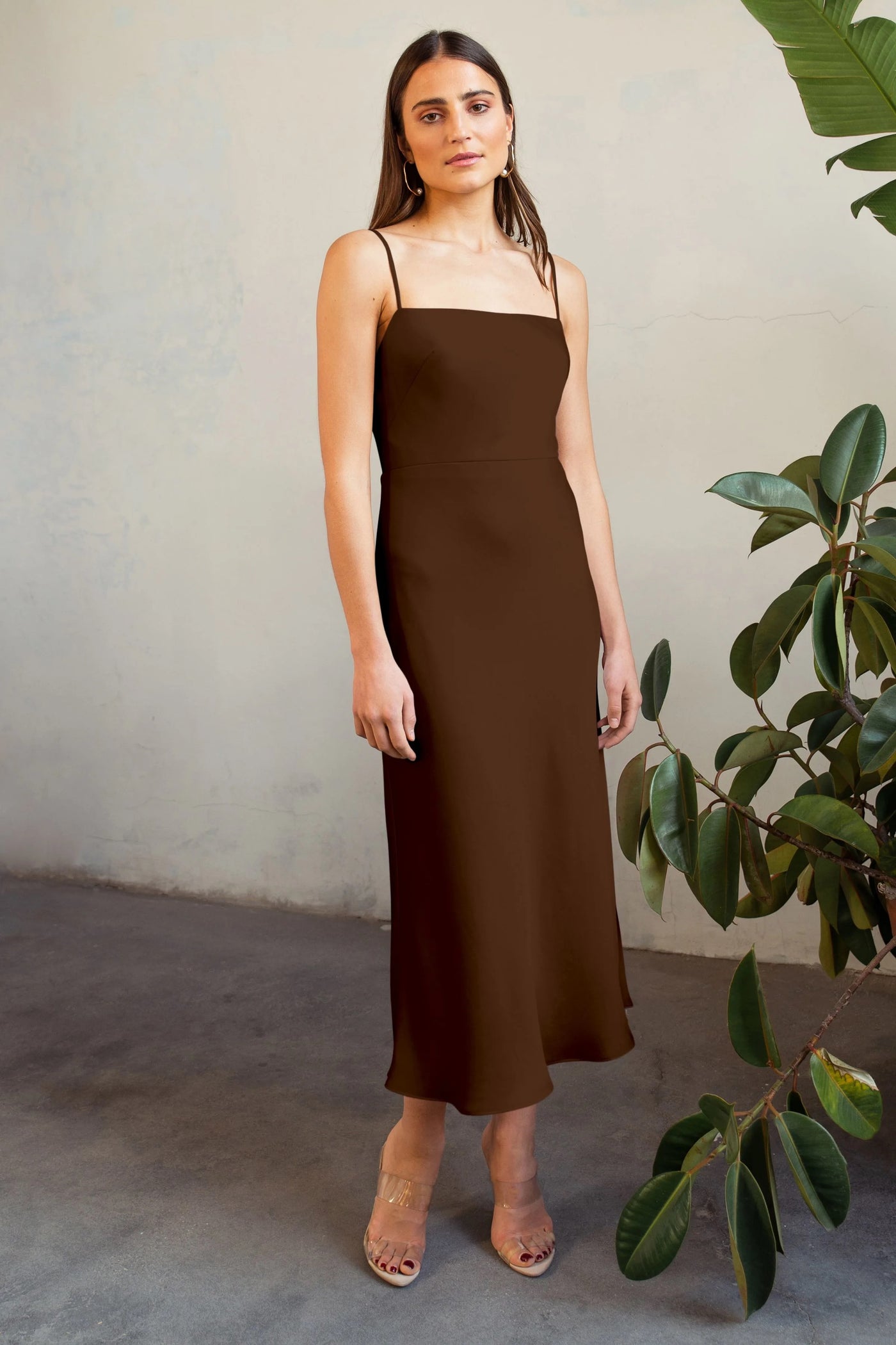 A woman in a brown satin midi dress stands near a plant, wearing clear heeled sandals, with a neutral expression. The dress she is wearing is the Claudia bridesmaid dress by Jenny Yoo from Bergamot Bridal.