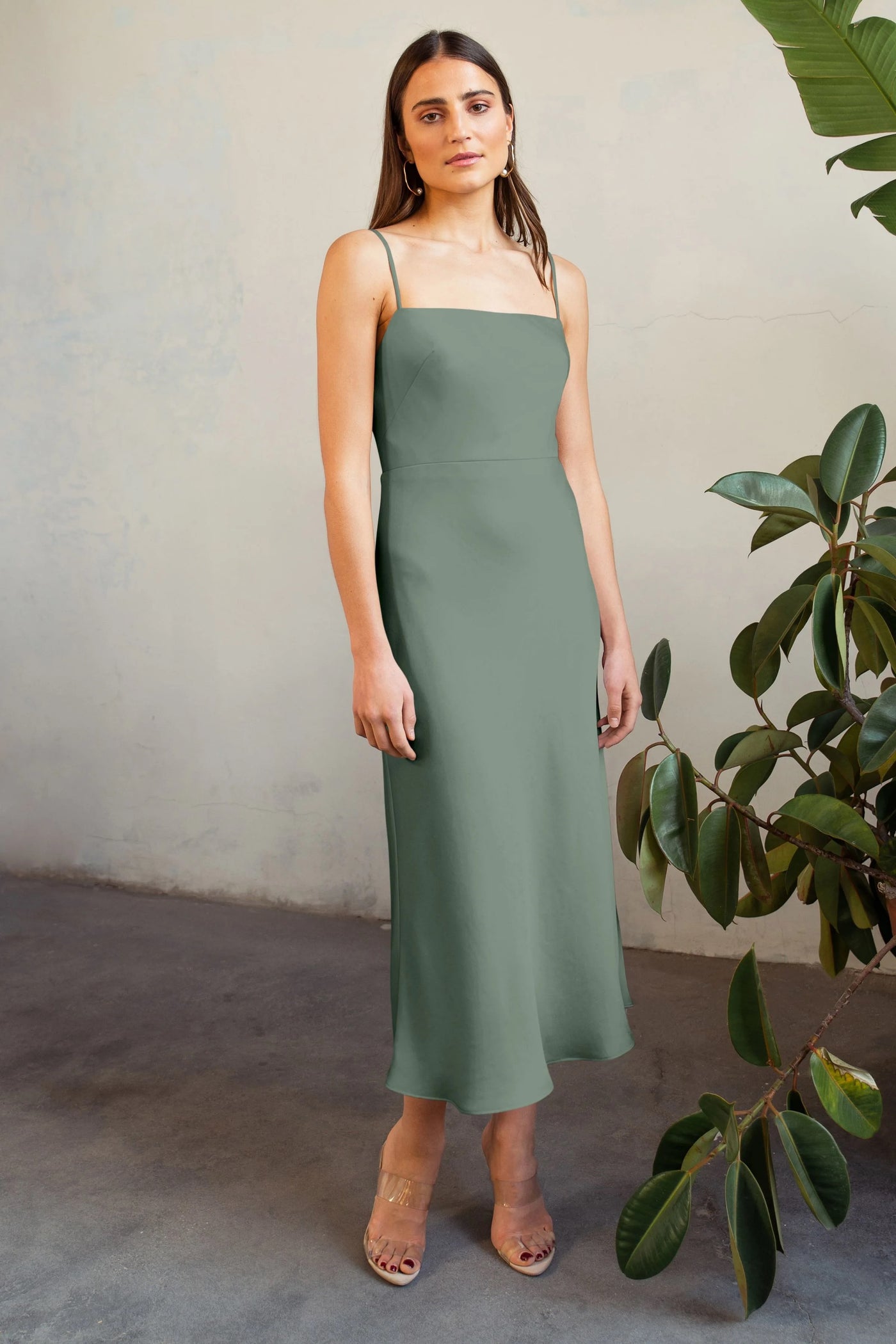 Woman in a Claudia - Bridesmaid Dress by Jenny Yoo in green satin with thin straps, standing by a plant, on a concrete floor against a textured wall.