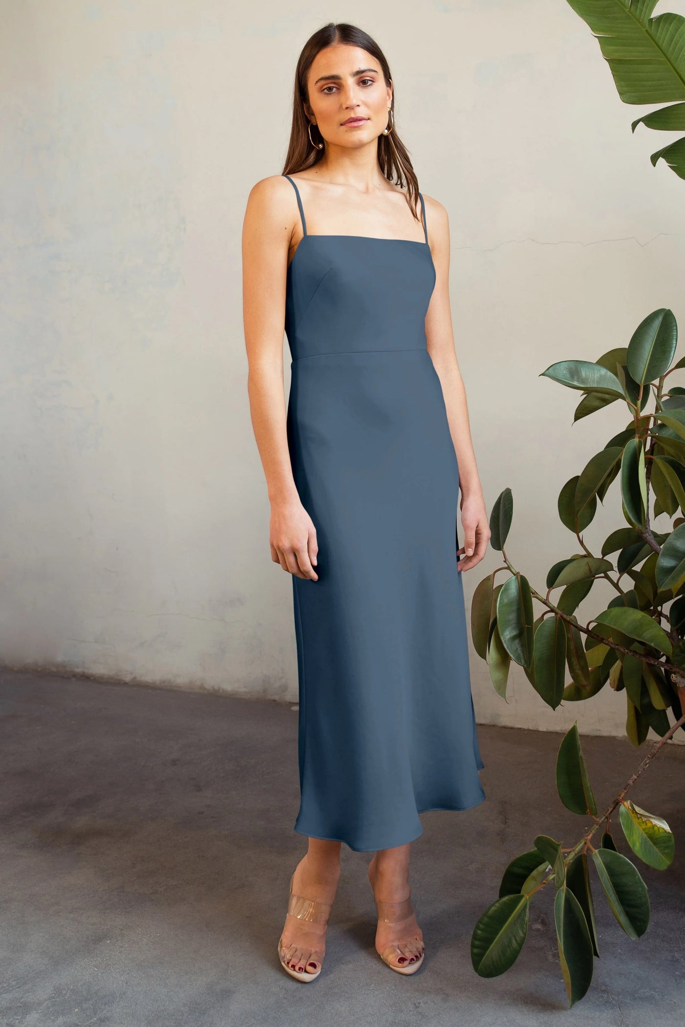 A woman stands in front of a plant-adorned wall wearing a Claudia bridesmaid dress by Jenny Yoo in slate blue satin and clear sandals from Bergamot Bridal.