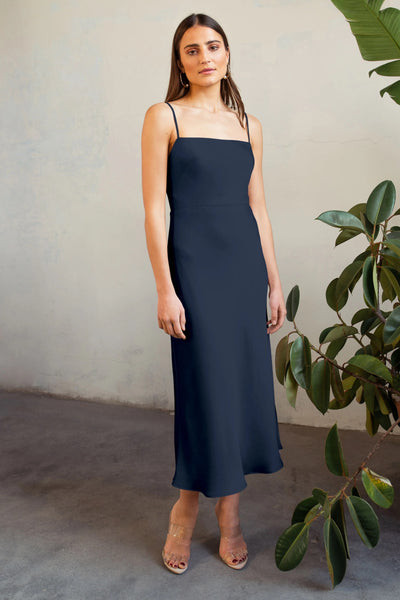 A woman in a navy blue satin midi Claudia - Bridesmaid Dress by Jenny Yoo stands near a foliage backdrop, wearing clear strap heels and looking at the camera.