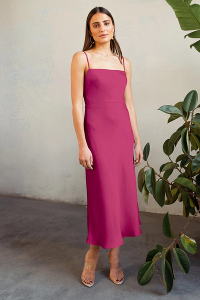 A woman stands against a textured wall, wearing a pink Claudia satin midi dress by Jenny Yoo and clear heels, beside a large green plant.