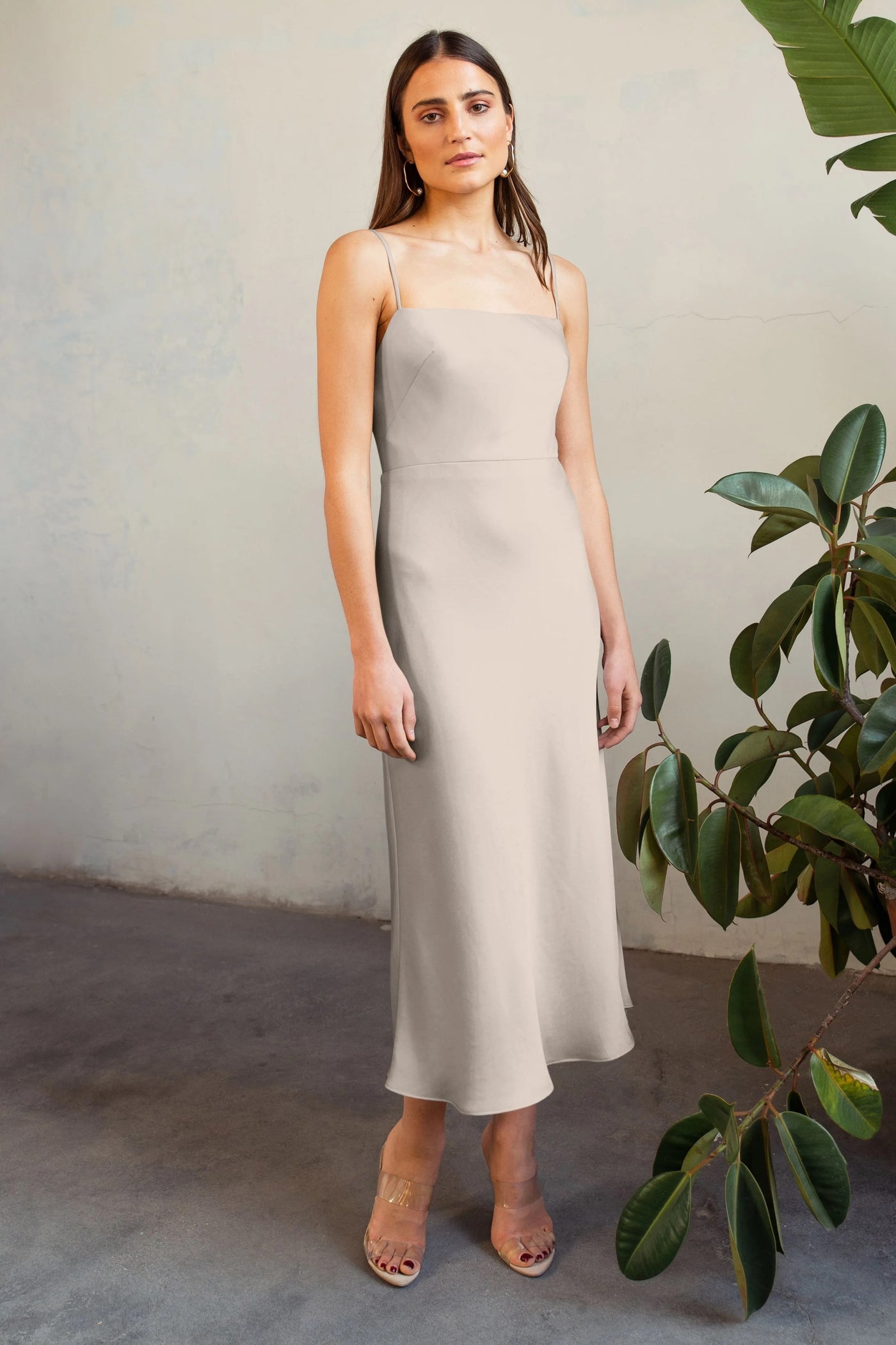 A woman in a beige satin midi dress with spaghetti straps and clear heels standing against a textured wall with a potted plant beside her, wearing the Claudia - Bridesmaid Dress by Jenny Yoo from Bergamot Bridal.