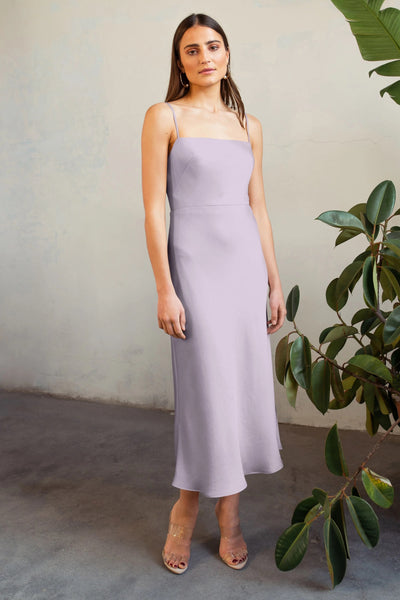 A woman stands in front of a textured wall, wearing a Claudia Bridesmaid Dress by Jenny Yoo with spaghetti straps, complemented by clear heeled sandals.