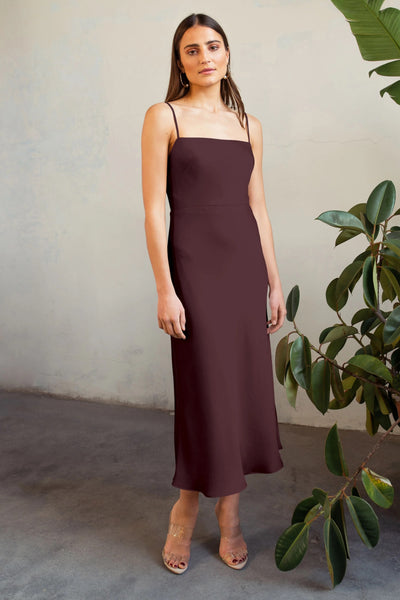 Woman in a maroon Claudia - Bridesmaid Dress by Jenny Yoo standing beside a green plant with a neutral expression.