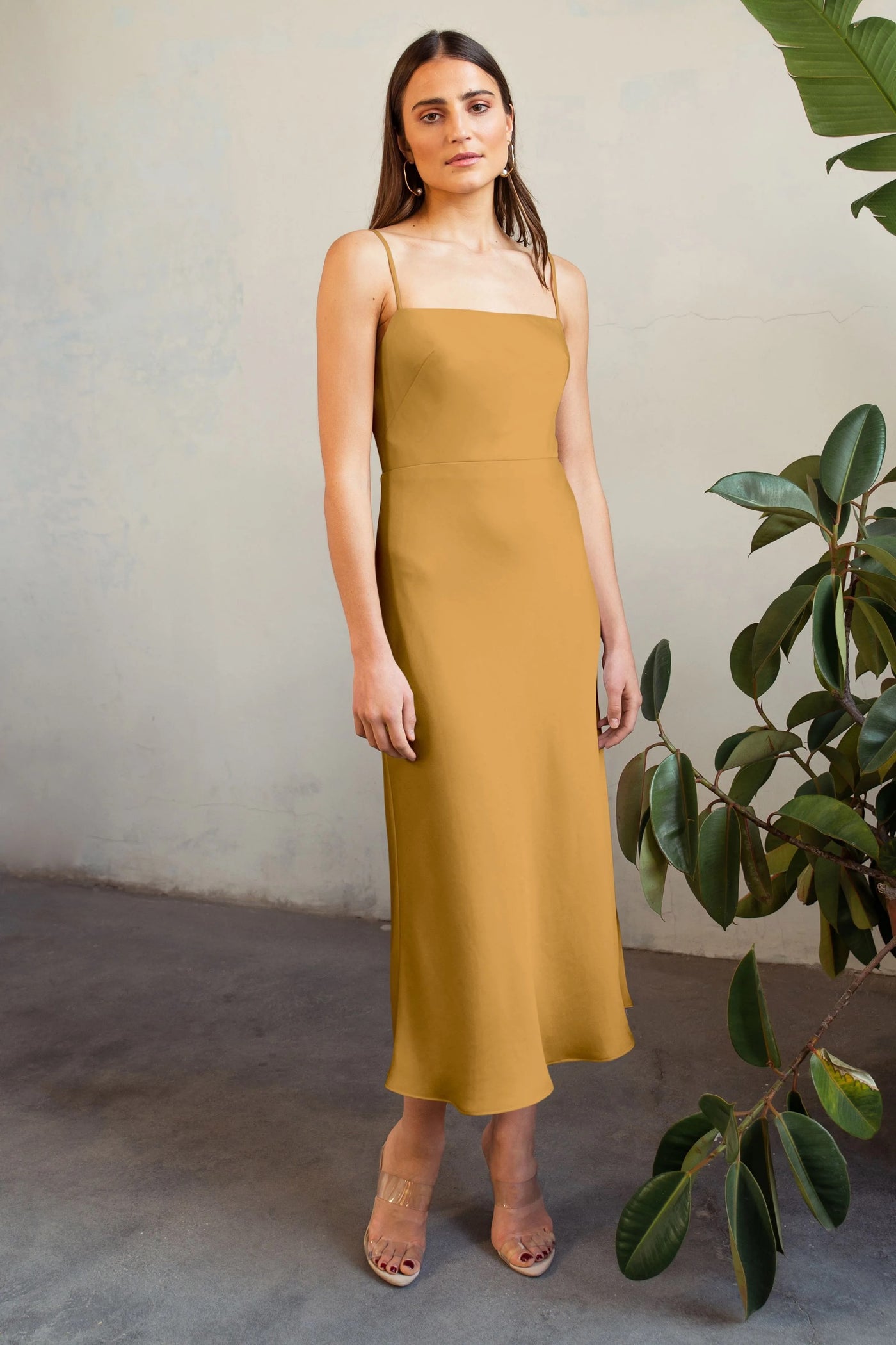 A woman in a Claudia - Bridesmaid Dress by Jenny Yoo with a straight neckline standing beside a plant, with a plain wall in the background.