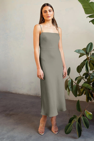 A woman standing in a plant-filled room wearing a sleeveless, olive green Claudia satin midi dress by Jenny Yoo and clear heeled sandals.