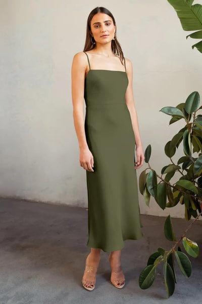 A woman in a green Claudia satin midi dress by Jenny Yoo and clear heels standing next to a potted plant.