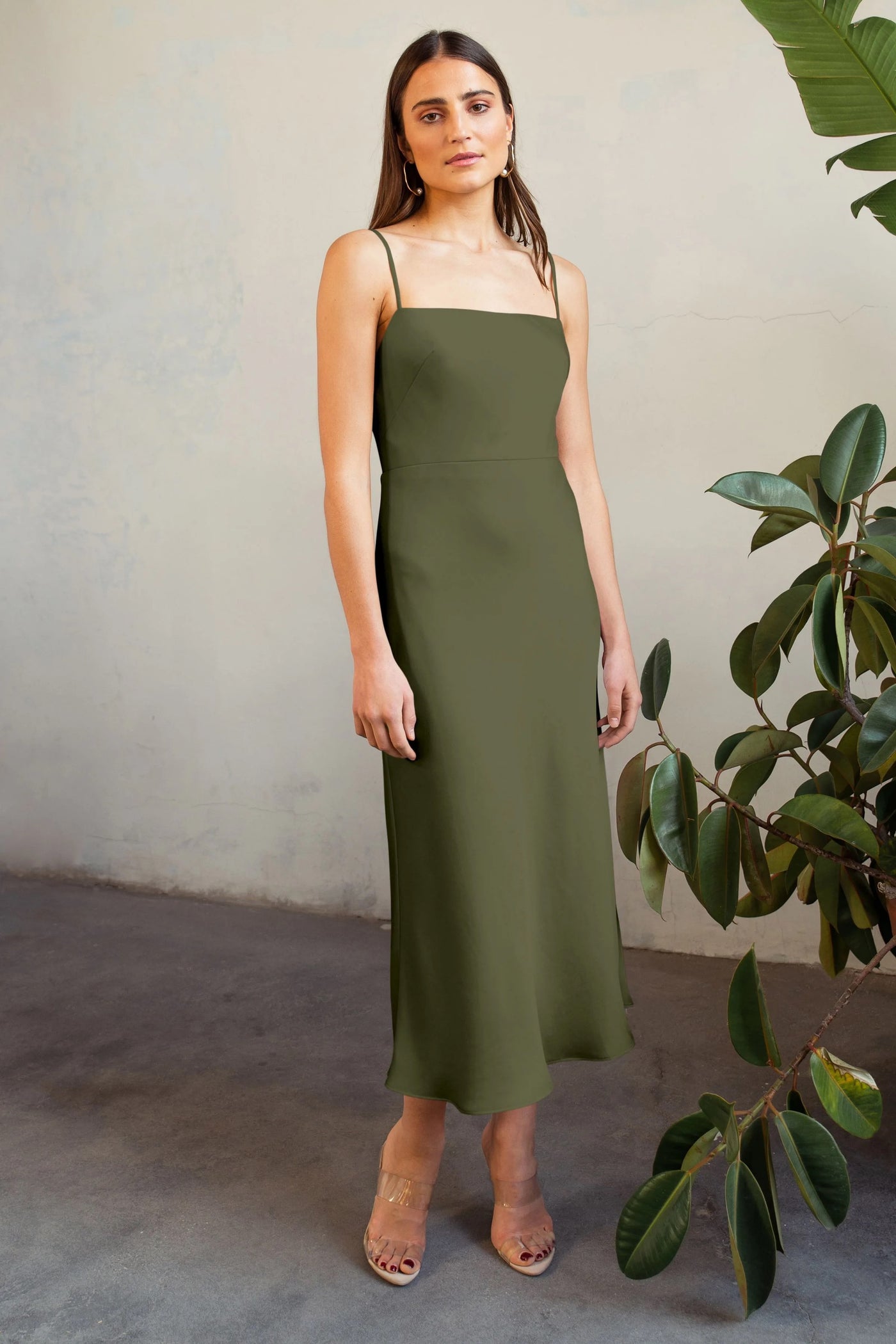 Woman in a green satin midi dress standing against a textured wall with a plant to her right wearing the Claudia Bridesmaid Dress by Jenny Yoo from Bergamot Bridal.