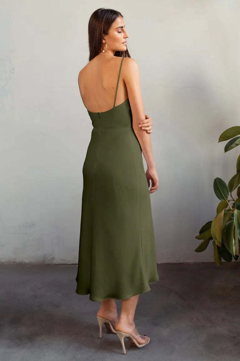 A woman from behind wearing the Claudia Bridesmaid Dress by Jenny Yoo in olive green satin with spaghetti straps and beige high heels, standing in front of a concrete wall with a potted plant to the side.