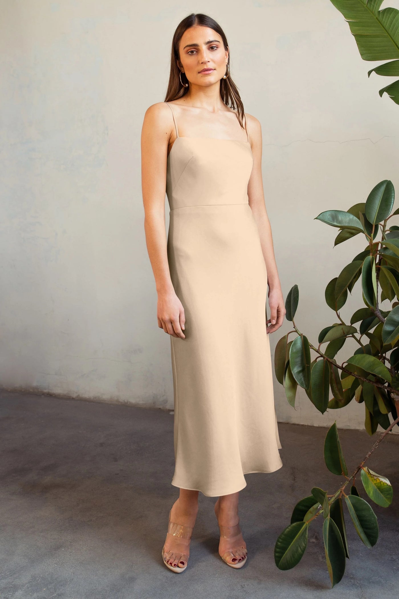 A woman in a beige Claudia satin midi dress by Jenny Yoo and clear heels stands beside a potted plant against a textured wall.