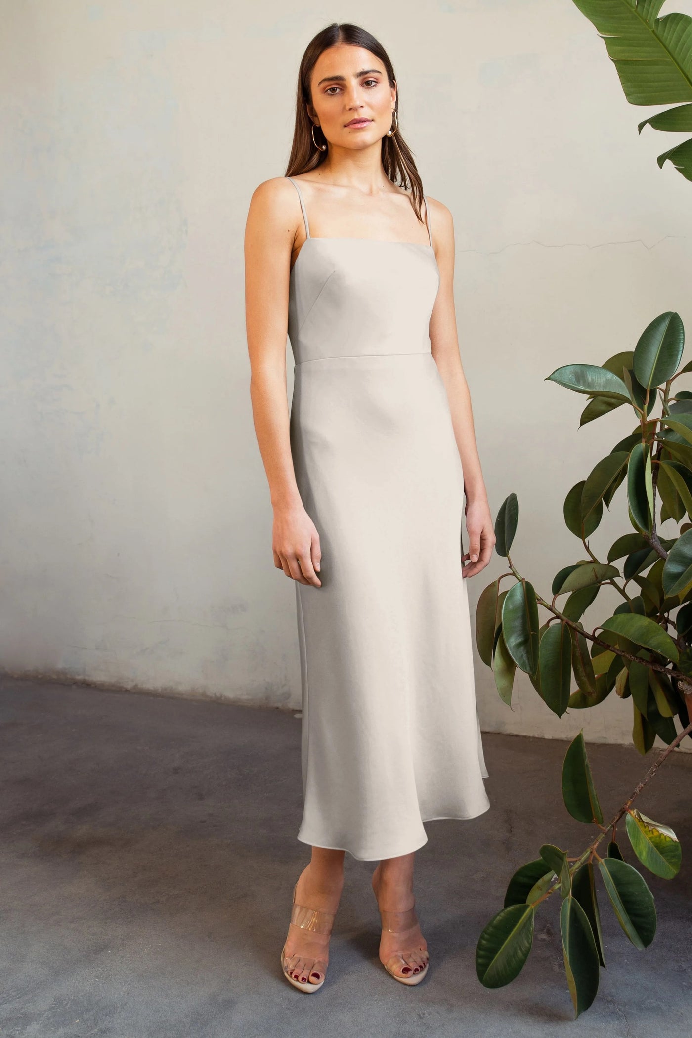 A woman in a light grey satin midi dress (Claudia - Bridesmaid Dress by Jenny Yoo) standing beside a green leafy plant, looking directly at the camera.