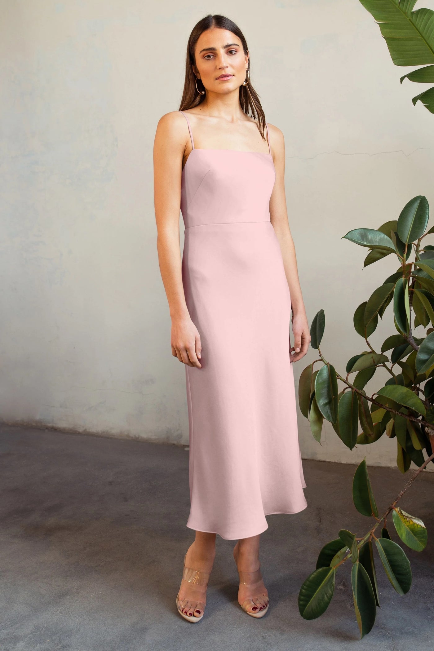 A woman in a sleeveless satin midi Claudia - Bridesmaid Dress by Jenny Yoo stands beside a large potted plant, looking directly at the camera.