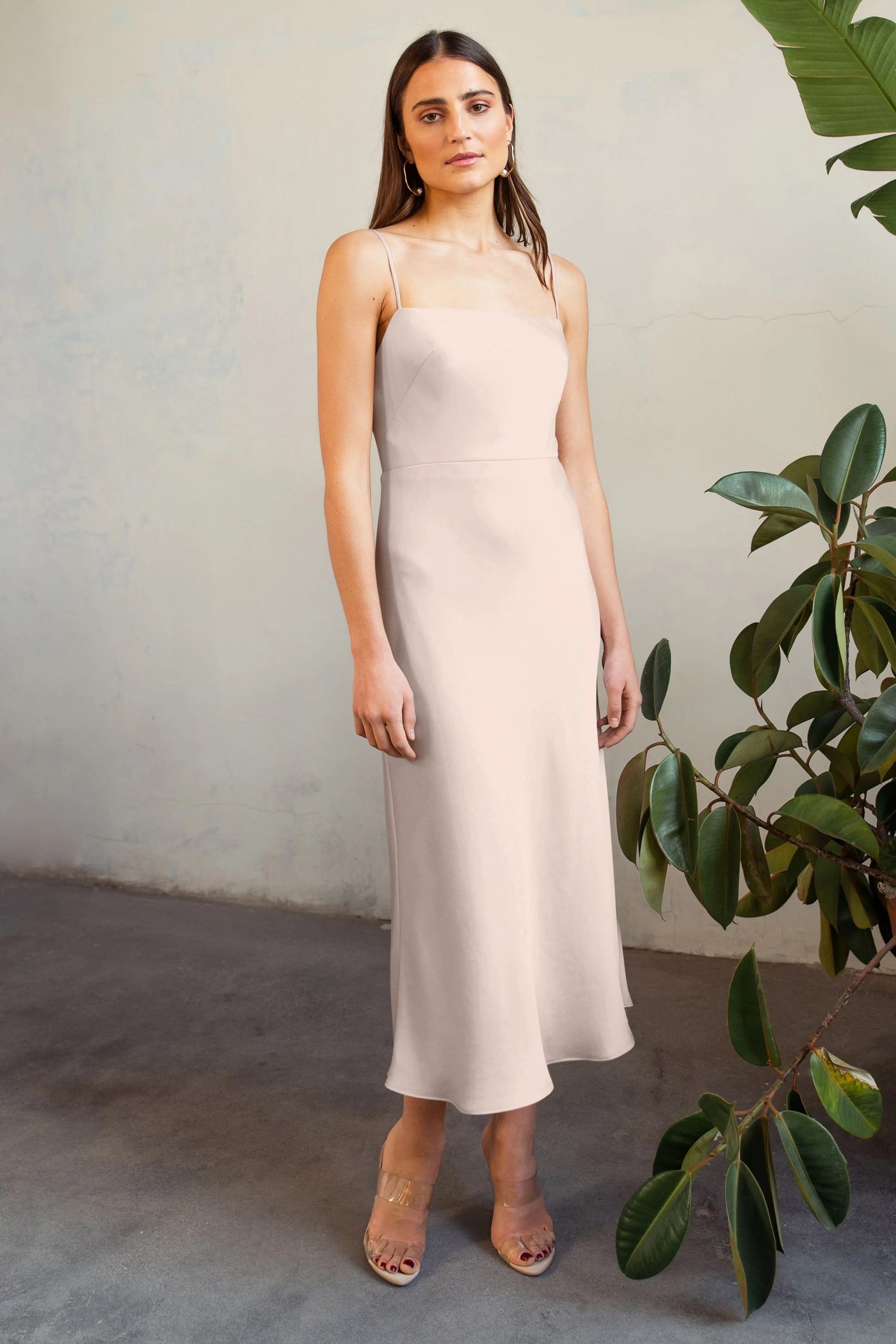 A woman in a Claudia - Bridesmaid Dress by Jenny Yoo by Bergamot Bridal and clear heels stands in front of a wall, with a potted plant to the side.