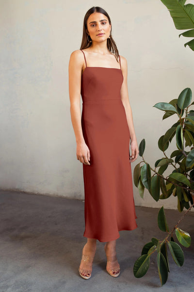 A woman in a sleeveless Jenny Yoo Claudia satin midi dress stands beside a potted plant, looking directly at the camera.