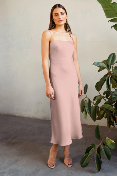 A woman in a pink Claudia - Bridesmaid Dress by Jenny Yoo stands next to a plant, with a textured wall in the background.