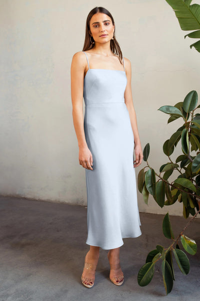 A woman in a light blue sleeveless Claudia satin midi dress by Jenny Yoo stands near a plant, looking at the camera with a neutral expression.