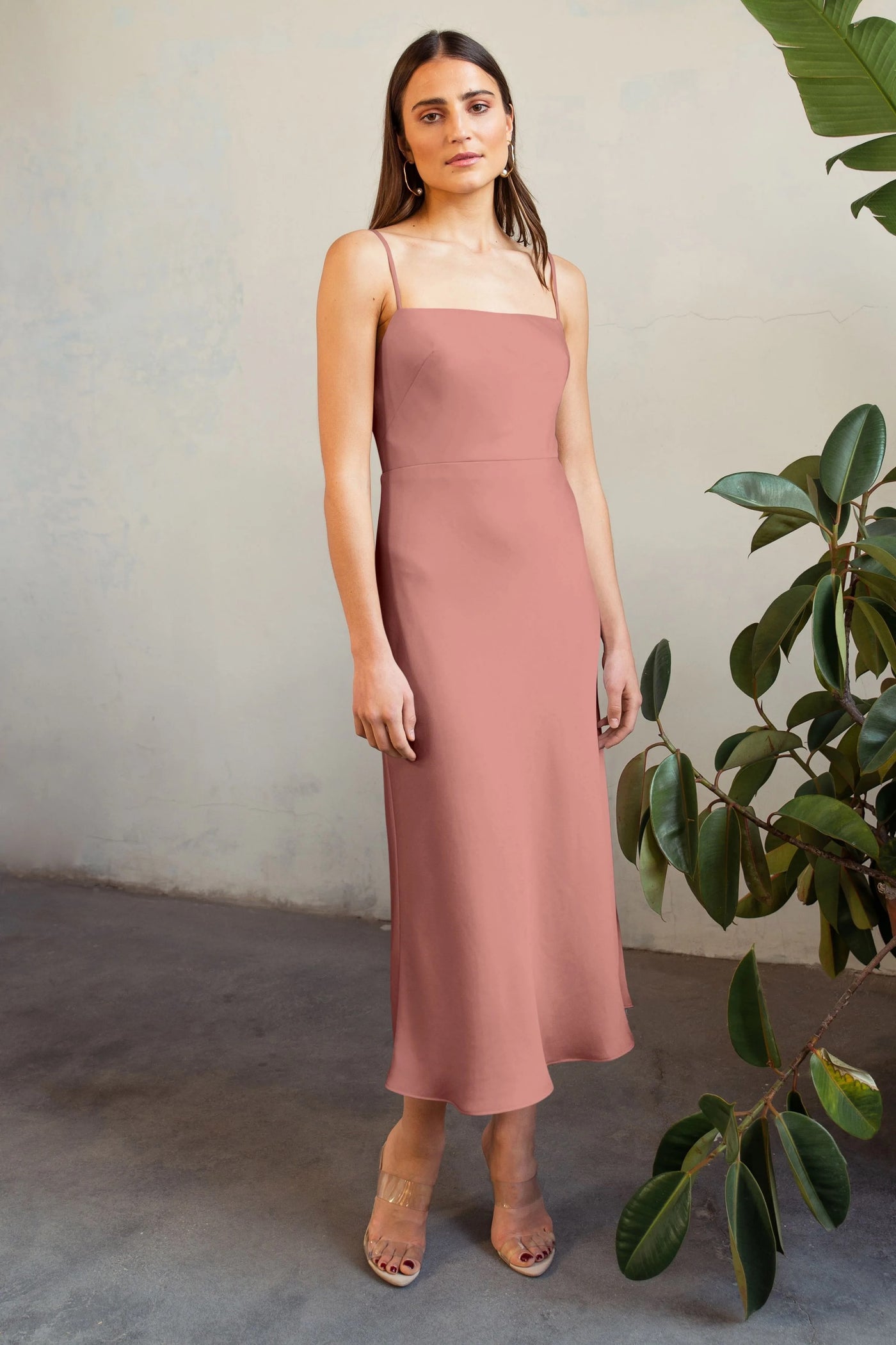 Woman in a rose-colored Claudia satin midi dress by Jenny Yoo standing against a textured wall with a potted plant nearby.