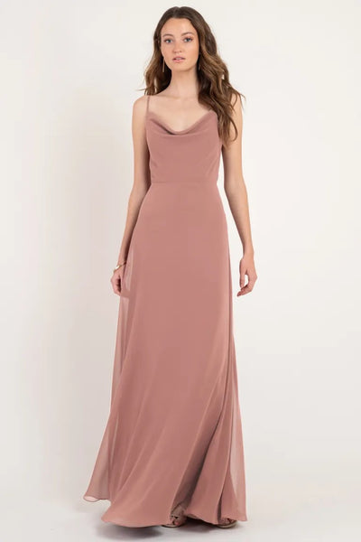 Woman in a long, sleeveless, mauve Colby - Jenny Yoo bridesmaid dress with a cowl neckline standing against a neutral background from Bergamot Bridal.