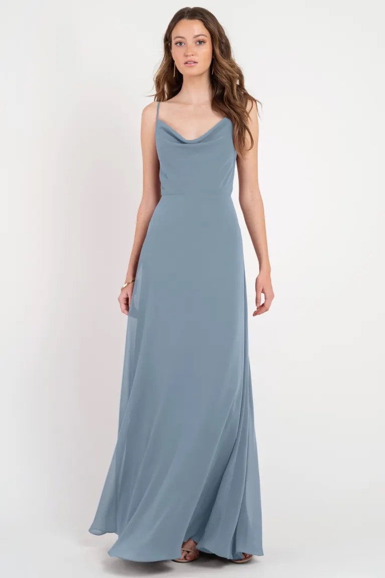 A woman standing in a Colby - Jenny Yoo Bridesmaid Dress from Bergamot Bridal, with a subtle cowl neckline and floor-length skirt.