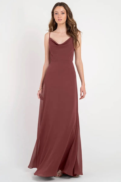 A woman in a sleeveless burgundy Colby - Jenny Yoo Bridesmaid Dress with a cowl neckline stands against a plain background from Bergamot Bridal.