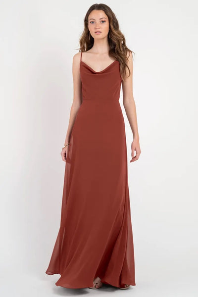 A woman in a long, rust-colored Colby - Jenny Yoo bridesmaid dress with a cowl neckline posing against a plain background from Bergamot Bridal.