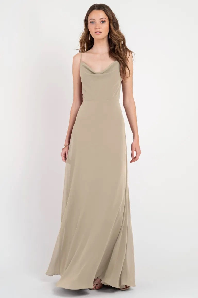 A woman standing in a neutral pose wearing an elegant, long beige slip dress with a cowl neckline, the Colby - Jenny Yoo Bridesmaid Dress from Bergamot Bridal.