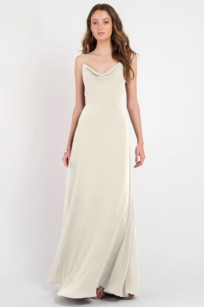 Woman modeling a floor-length cream slip dress with thin straps and a cowl neckline, the Colby by Jenny Yoo bridesmaid dress from Bergamot Bridal.
