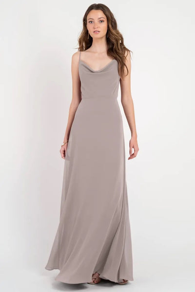 A woman wearing a long, elegant taupe slip dress with a cowl neckline, the Colby - Jenny Yoo Bridesmaid Dress from Bergamot Bridal.