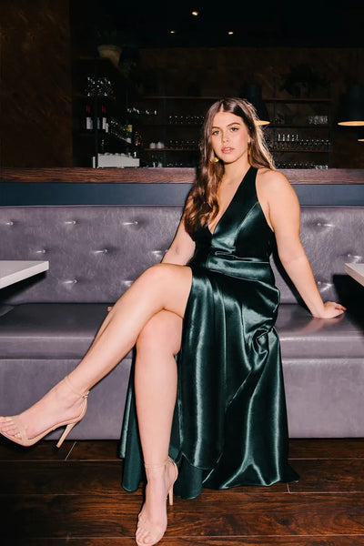 A woman wearing the Corinne Bridesmaid Dress by Bergamot Bridal, crafted from dark green satin, sits elegantly on a gray booth seat in a dimly lit setting, her leg gracefully crossed over the other.