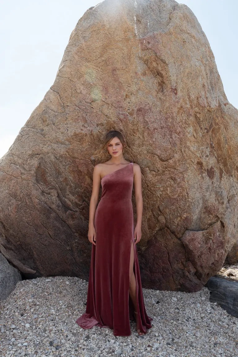 Woman in an elegant one shoulder red Cybill - Bridesmaid Dress by Jenny Yoo made of stretch velvet, standing in front of a large boulder.