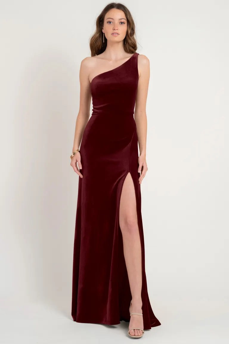 A woman in a Cybill bridesmaid dress by Jenny Yoo in stretch velvet, one-shoulder style, burgundy color, with a high slit, available in store sample size 12 at Bergamot Bridal.