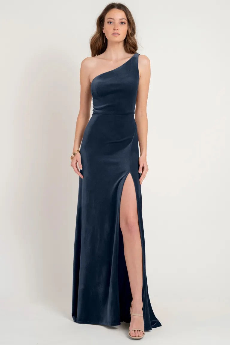 A woman wearing a Cybill bridesmaid dress by Jenny Yoo, a one-shoulder, navy blue evening gown made of stretch velvet with a high slit from Bergamot Bridal.