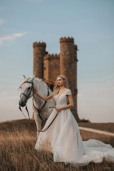 A woman in a Bergamot Bridal Allure Disney Wedding Dress - Rapunzel - Off The Rack stands with a white horse in front of an old castle tower, embarking on her wedding adventure.