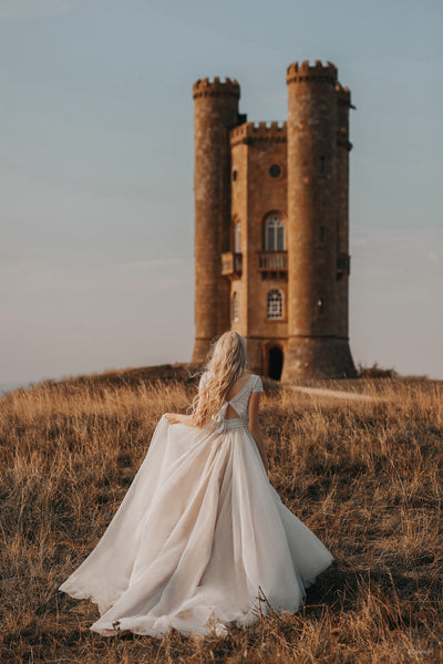 A woman in a Bergamot Bridal Allure Disney Wedding Dress - Rapunzel - Off The Rack standing in a field gazing at an old tower.