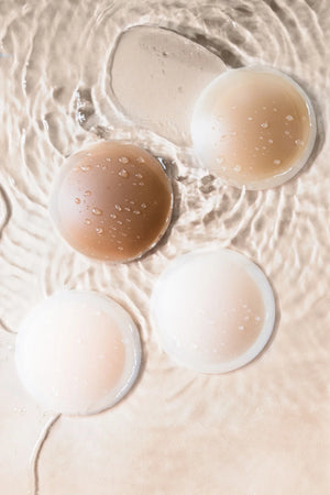 Four circular Threads Invisible Reusable Nipple Stickies by Bergamot Bridal, featuring seamless tapered edges in light to dark brown shades, rest on a rippled water surface. Water droplets enhance their smooth design.