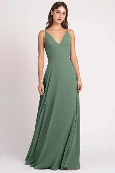 A woman in a green, v-neck, floor-length Dani - Bridesmaid Dress by Jenny Yoo with an a-line silhouette standing against a neutral background from Bergamot Bridal.