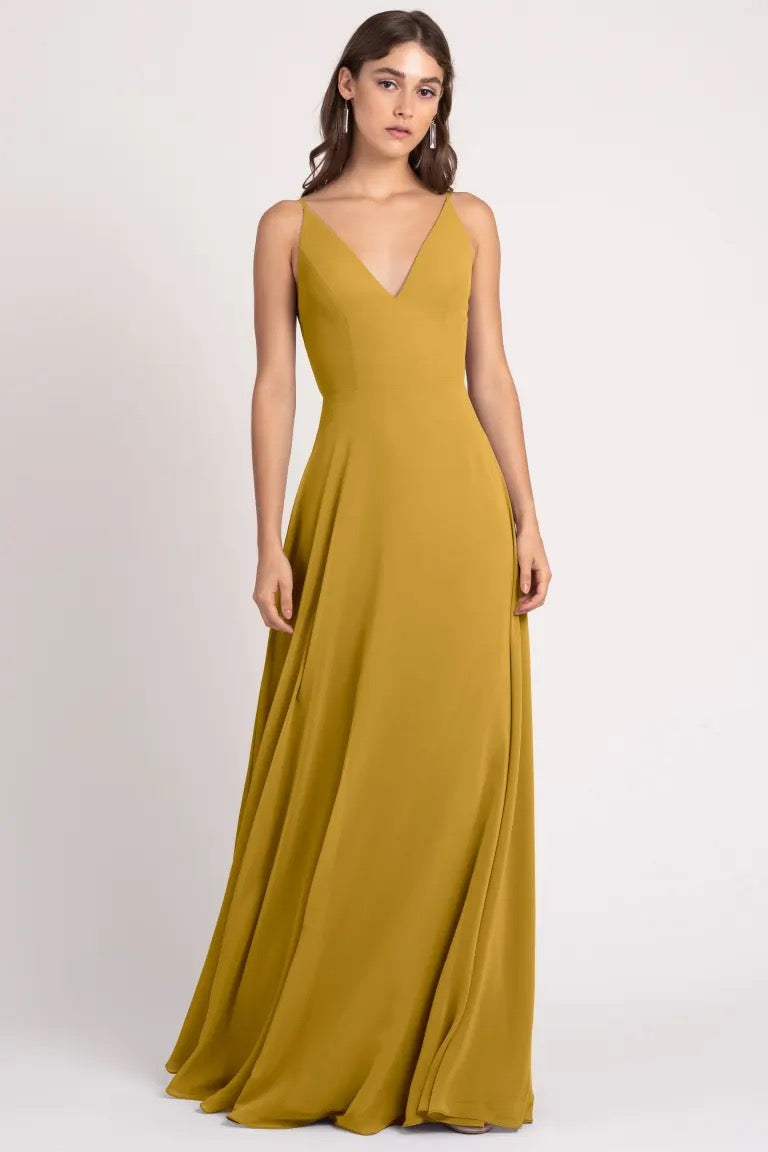 A woman in a long mustard evening gown with a deep v-neckline and sharp tailoring poses against a neutral backdrop. The Dani Bridesmaid Dress by Jenny Yoo from Bergamot Bridal.